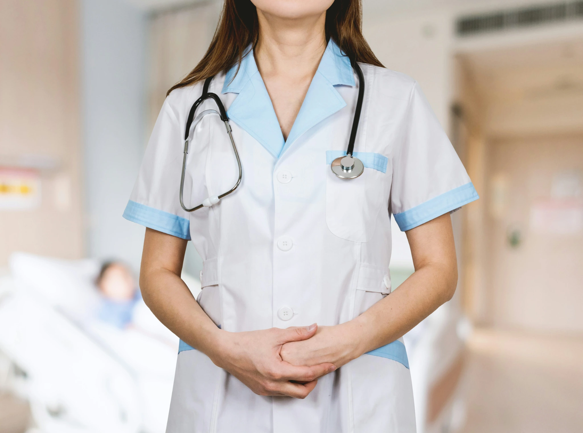 Healthcare professional in a white uniform with a stethoscope standing with hands clasped, with a hospital bed in the background.