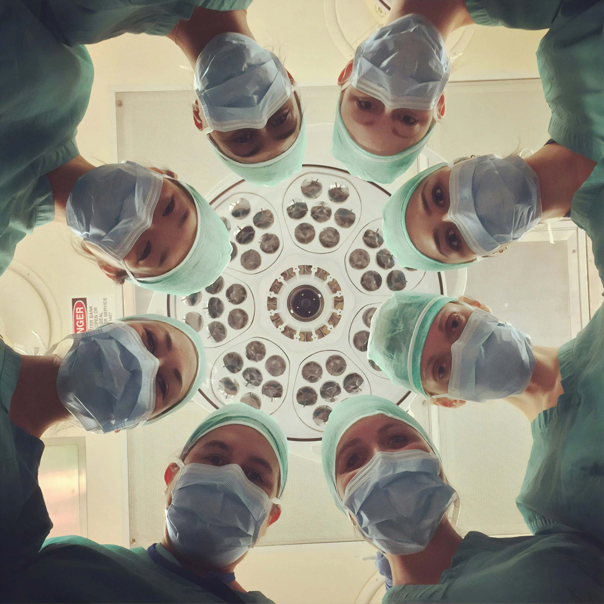 Group of surgeons wearing surgical masks and caps looking down at the camera under an operating theatre light.