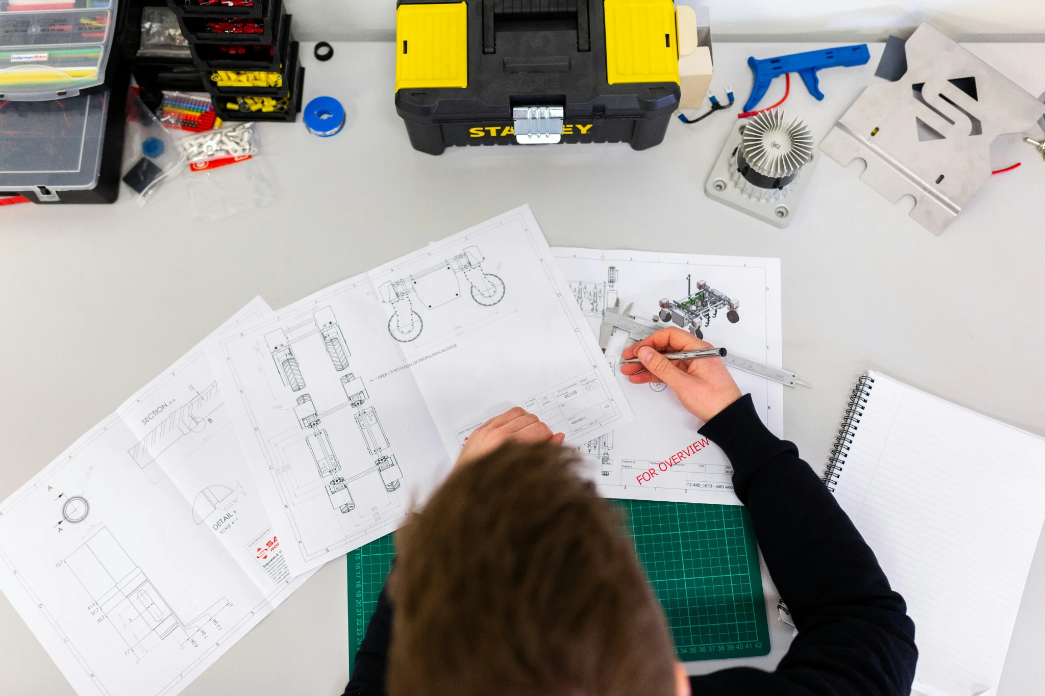 Top-down view of a person writing in a notebook surrounded by technical drawings and tools.